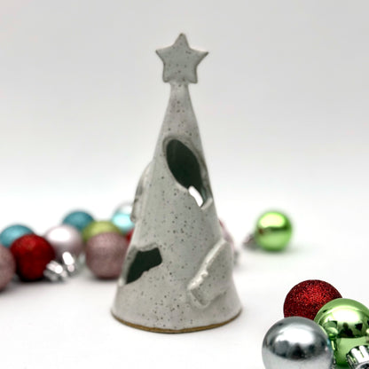 A white speckled ceramic tree luminary with cutout holiday light shapes and a star on top, surrounded by small colorful ornaments.