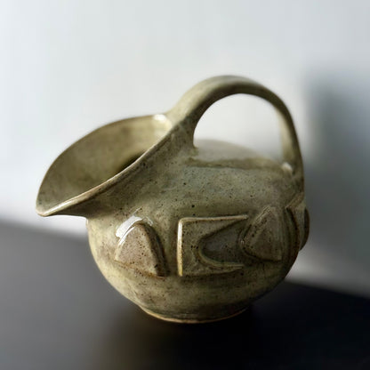 Small handmade stoneware jug with a rounded body, arched handle, curved spout, and raised mid-century geometric design, finished in a warm cream glaze.