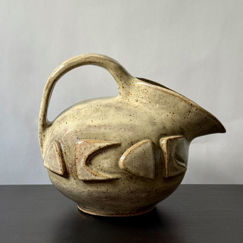 Small handmade stoneware jug with a rounded body, arched handle, curved spout, and raised mid-century geometric design, finished in a warm cream glaze.