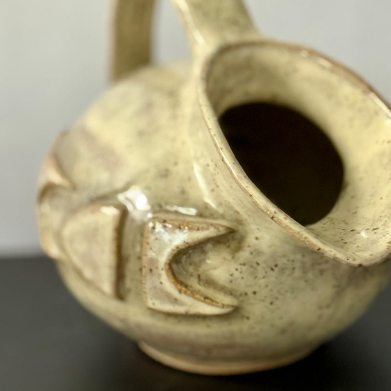 Small handmade stoneware jug with a rounded body, arched handle, curved spout, and raised mid-century geometric design, finished in a warm cream glaze.