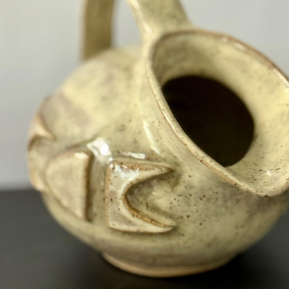 Small handmade stoneware jug with a rounded body, arched handle, curved spout, and raised mid-century geometric design, finished in a warm cream glaze.