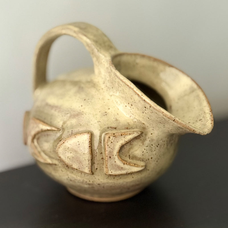 Small handmade stoneware jug with a rounded body, arched handle, curved spout, and raised mid-century geometric design, finished in a warm cream glaze.