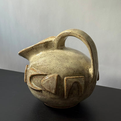 Small handmade stoneware jug with a rounded body, arched handle, curved spout, and raised mid-century geometric design, finished in a warm cream glaze.