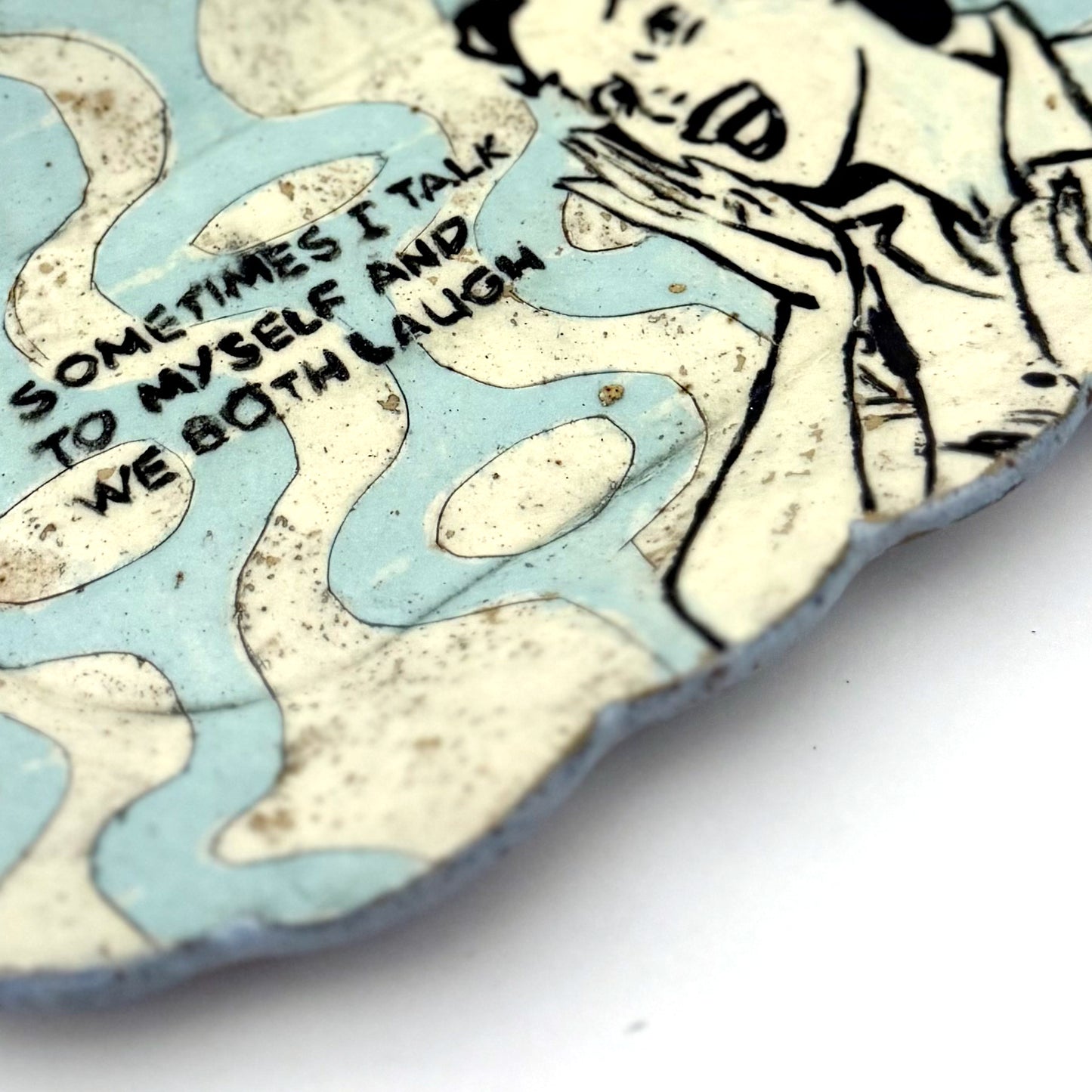 Close-up of a handmade scalloped-edge ceramic plate with a retro illustrated woman, blue and cream mid-century geometric pattern, and the phrase “Sometimes I talk to myself and we both laugh.”