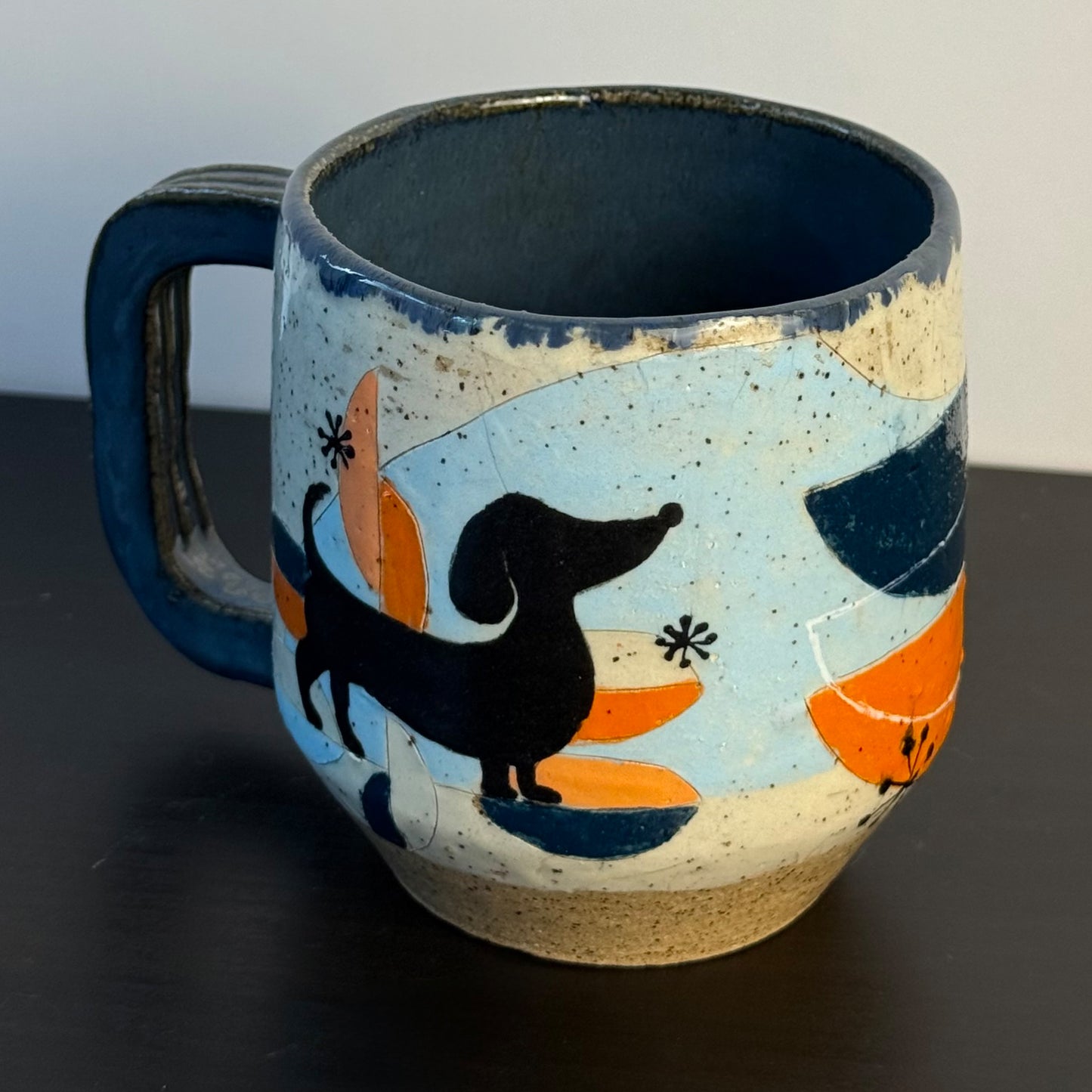 A handmade ceramic mug with a dark blue handle and interior, decorated with a black dachshund silhouette and orange and light blue abstract shapes on a speckled cream and blue glaze.