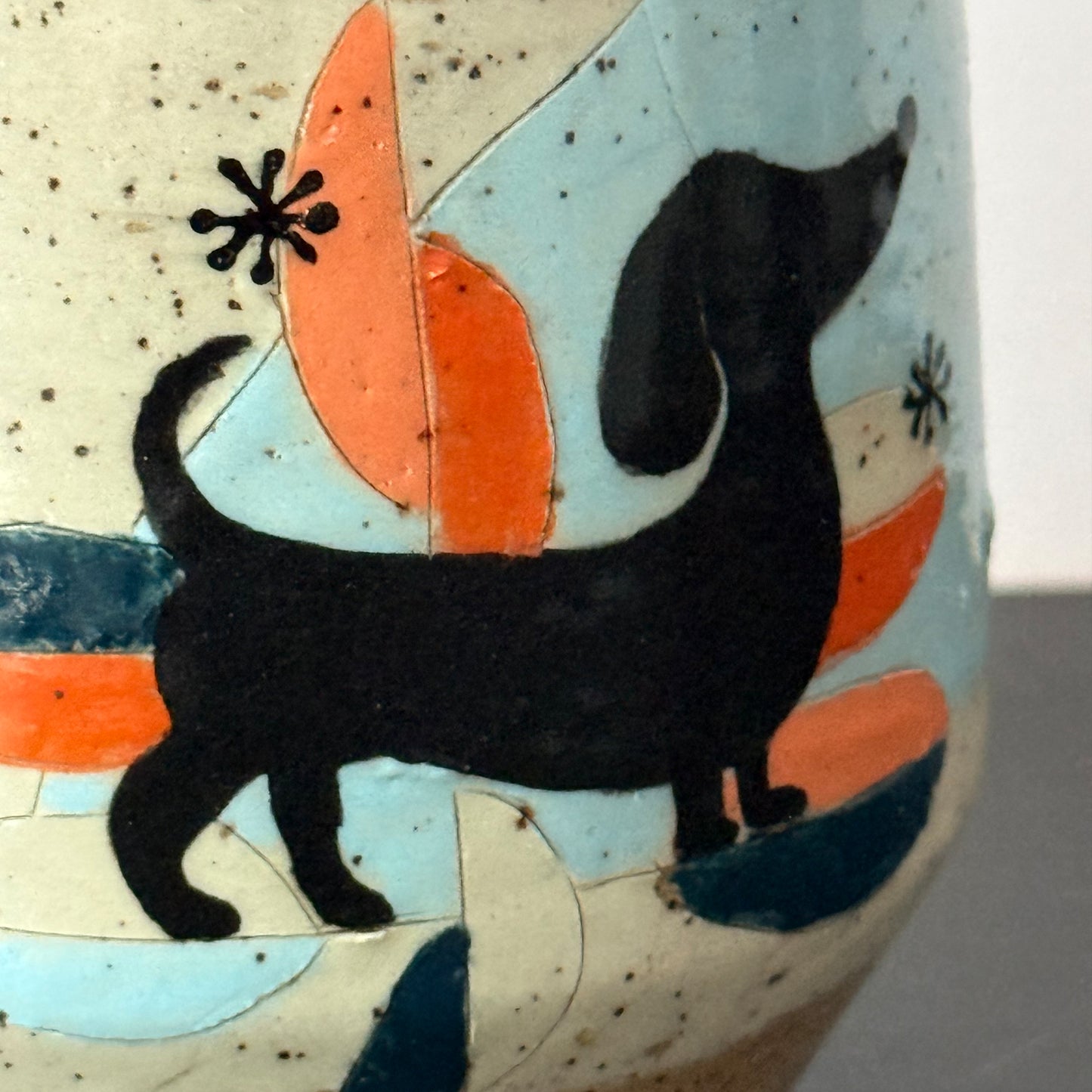 Close-up of a ceramic mug featuring a black dachshund silhouette over abstract orange, light blue, and dark blue shapes on a speckled cream background.