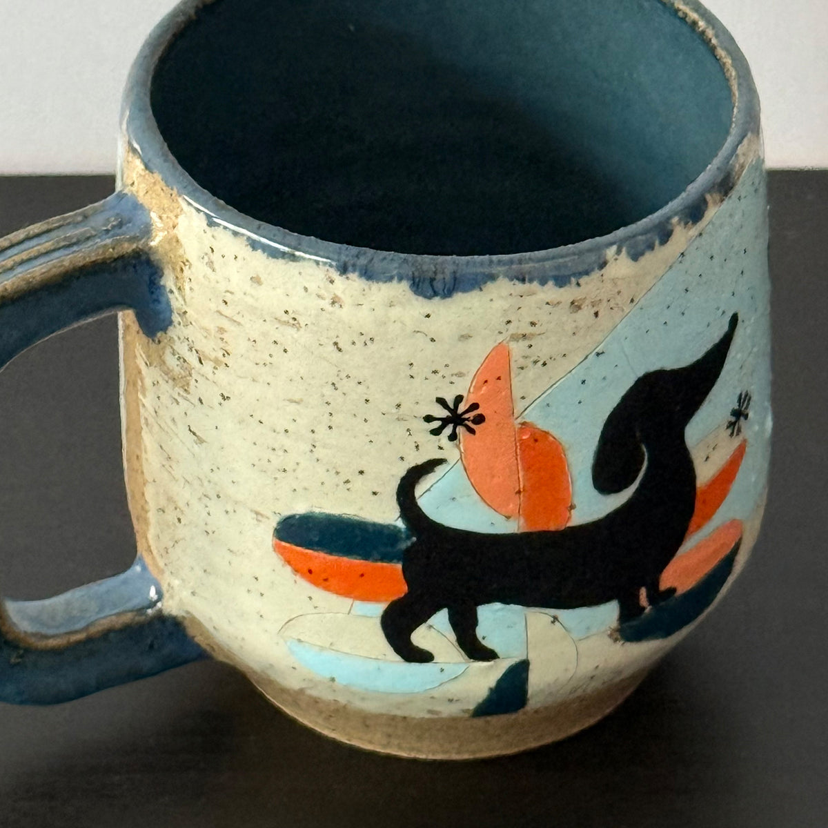 A handmade ceramic mug with a speckled cream exterior, a dark blue glazed interior and handle, and a colorful hand-painted design featuring a black dachshund, orange shapes, and light blue accents.