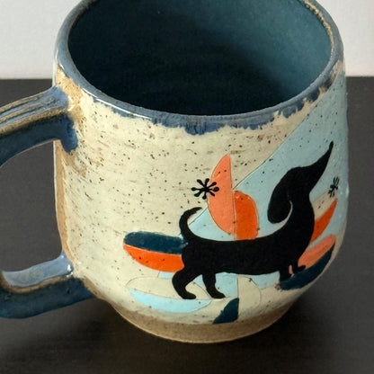 A handmade ceramic mug with a speckled cream exterior, a dark blue glazed interior and handle, and a colorful hand-painted design featuring a black dachshund, orange shapes, and light blue accents.