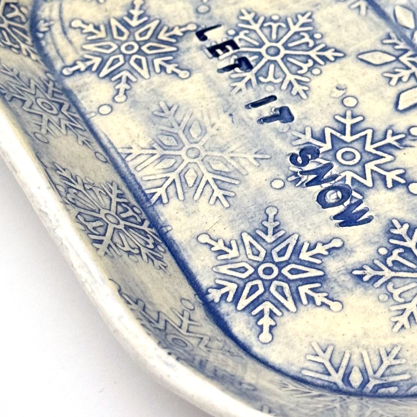 Close-up of an oval blue-and-white ceramic dish embossed with snowflakes, with “LET IT SNOW” stamped in the center, styled beside frosted pinecones.