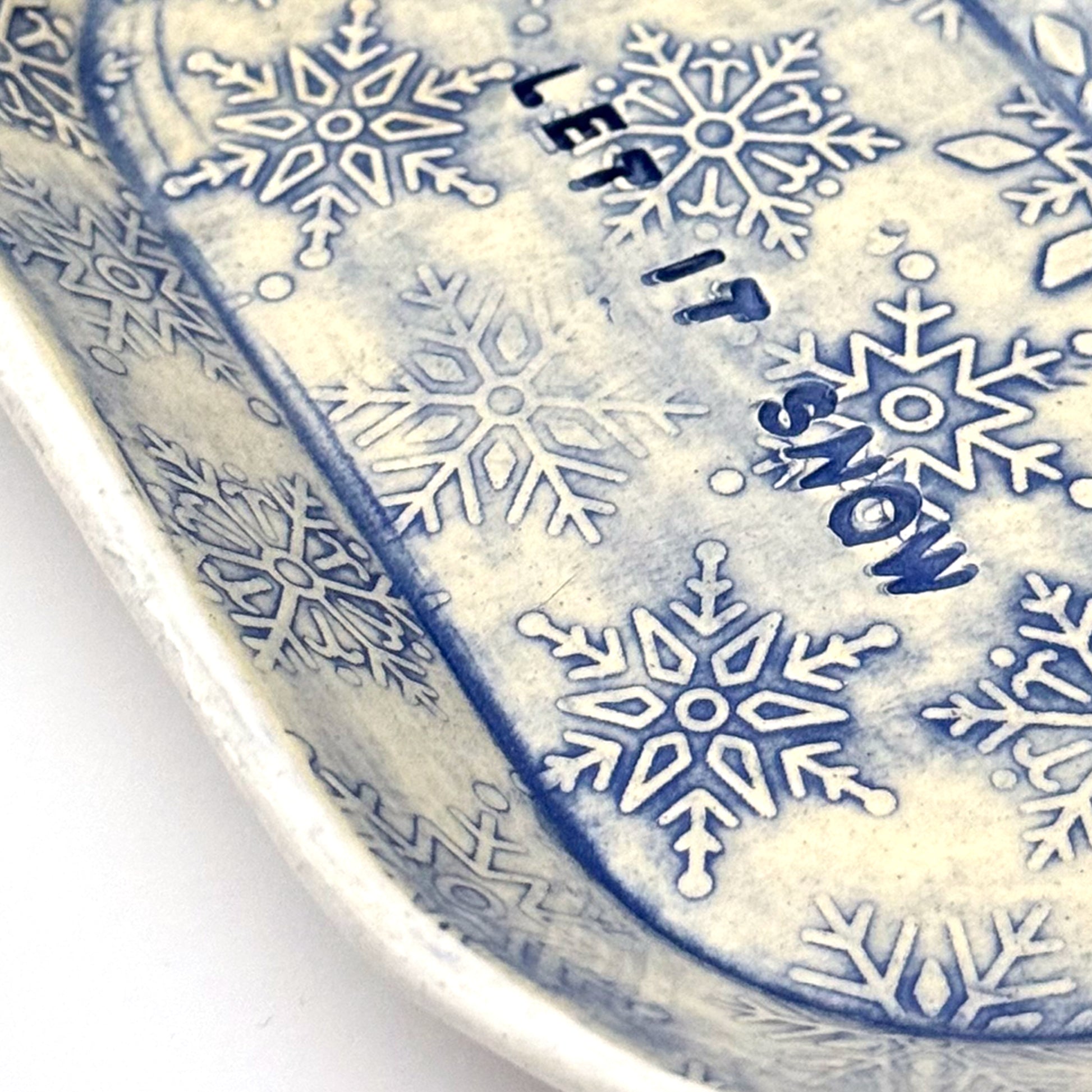 Close-up of an oval blue-and-white ceramic dish embossed with snowflakes, with “LET IT SNOW” stamped in the center, styled beside frosted pinecones.