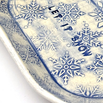 Close-up of an oval blue-and-white ceramic dish embossed with snowflakes, with “LET IT SNOW” stamped in the center, styled beside frosted pinecones.
