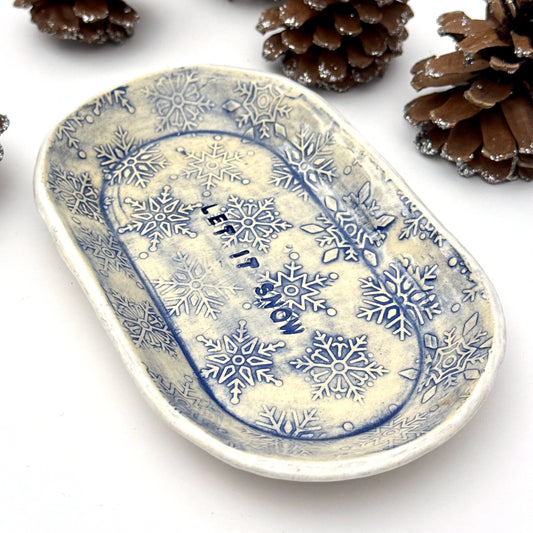 An oval blue-and-white ceramic dish embossed with snowflakes, with “LET IT SNOW” stamped in the center, styled beside frosted pinecones.