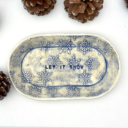 An oval blue-and-white ceramic dish embossed with snowflakes, with “LET IT SNOW” stamped in the center, styled beside frosted pinecones.
