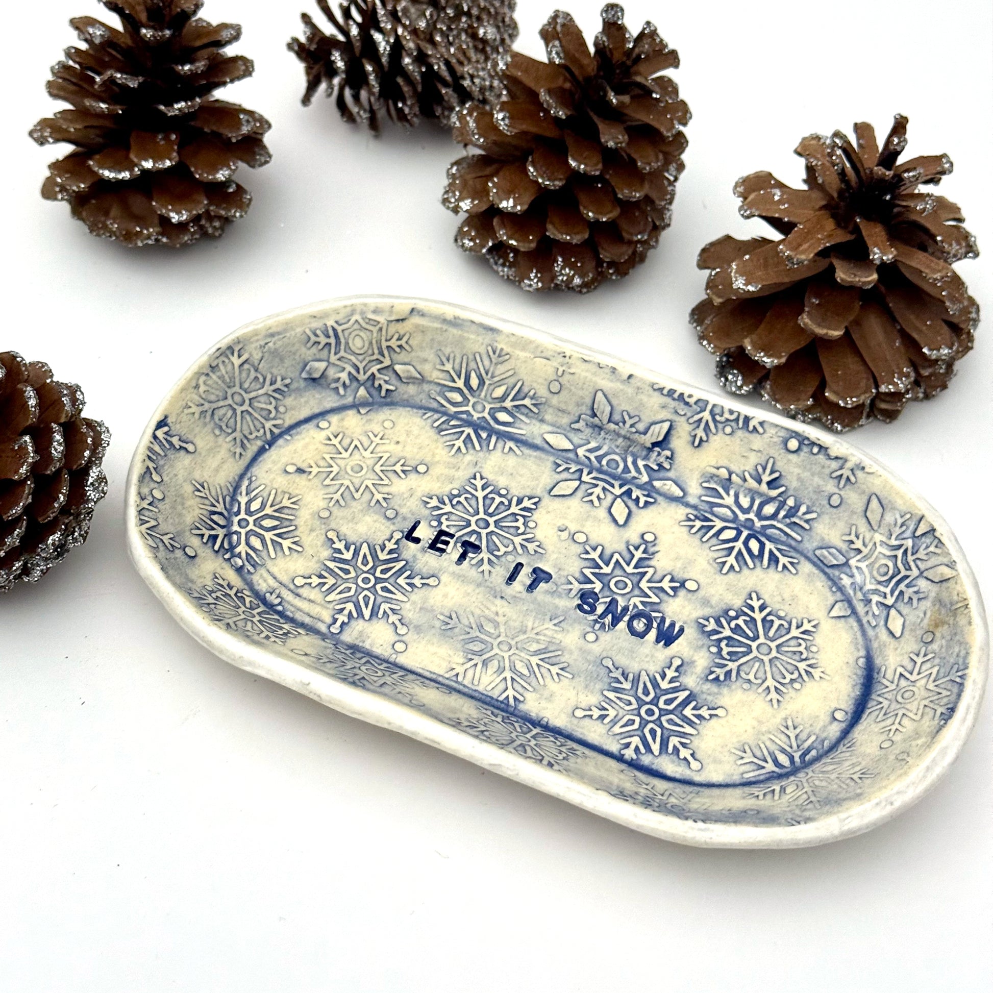 An oval blue-and-white ceramic dish embossed with snowflakes, with “LET IT SNOW” stamped in the center, styled beside frosted pinecones.