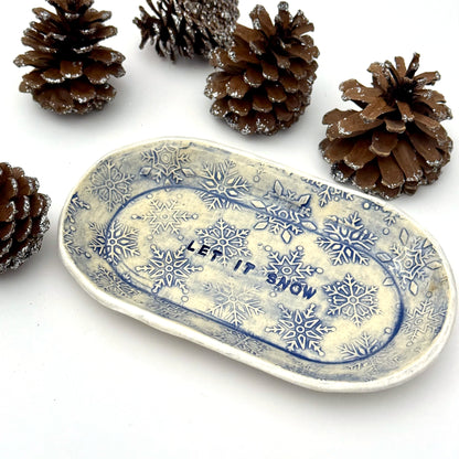 An oval blue-and-white ceramic dish embossed with snowflakes, with “LET IT SNOW” stamped in the center, styled beside frosted pinecones.