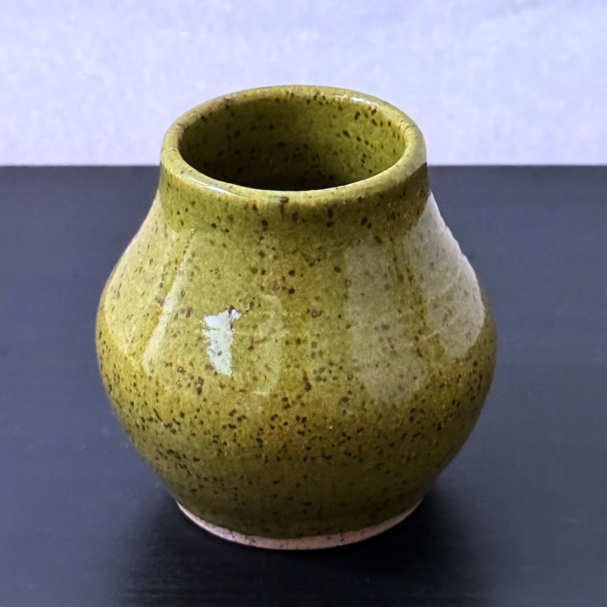 A small ceramic bud vase with a rounded body and flared rim, covered in a speckled olive green glaze, set against a dark surface and light background.