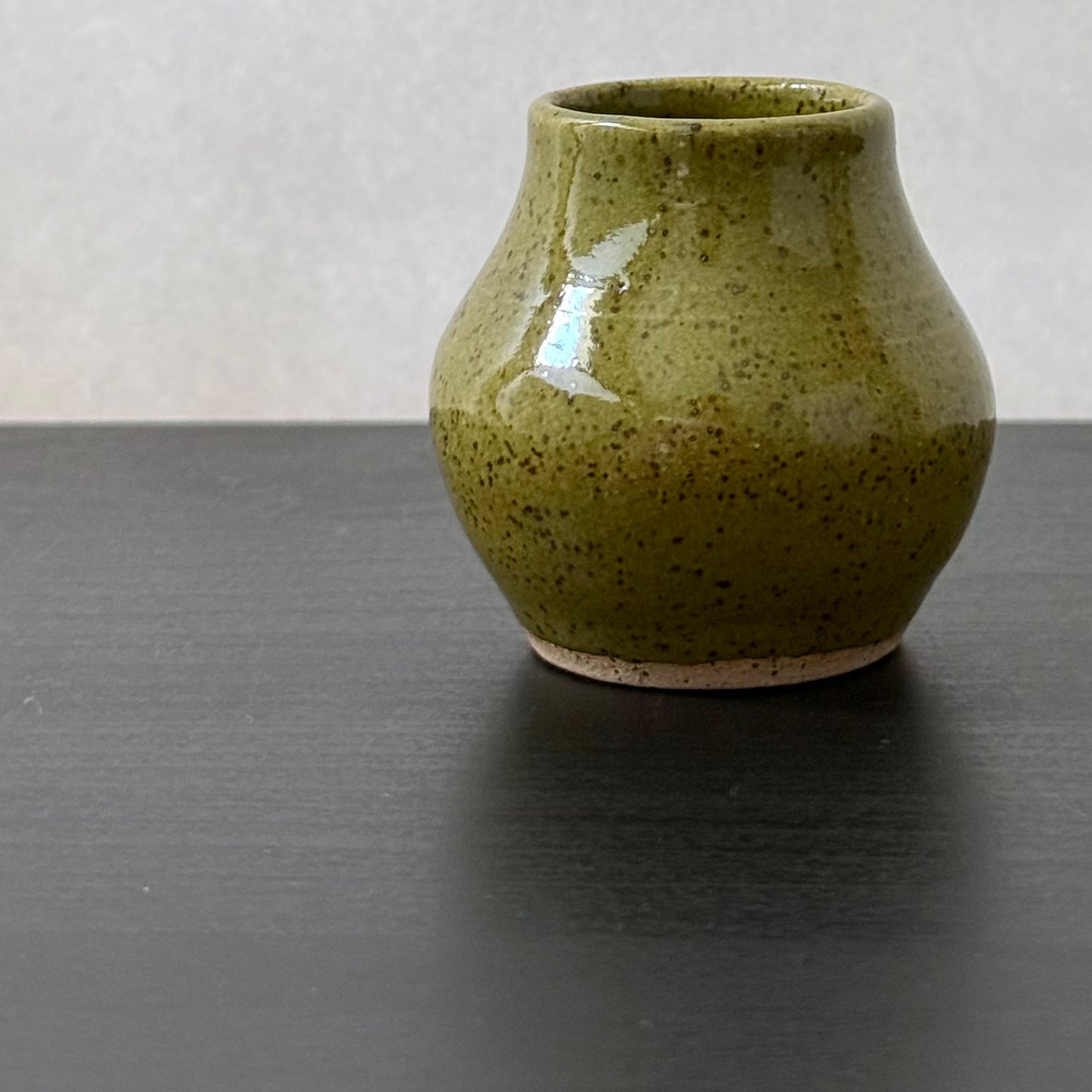 A small handmade ceramic vase with a rounded body, a flared rim, and a speckled olive green glaze, sitting on a dark wooden surface against a light gray background.