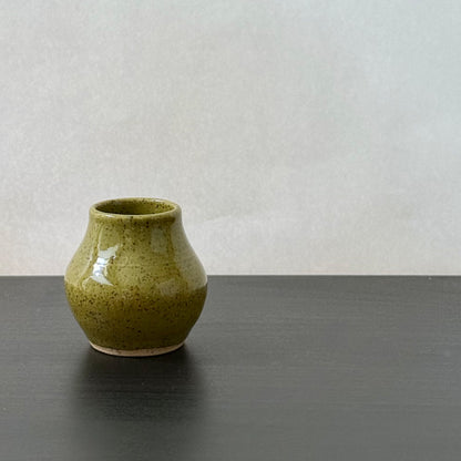 A small handmade ceramic vase with a speckled olive green glaze, sitting on a dark gray surface against a light gray background.