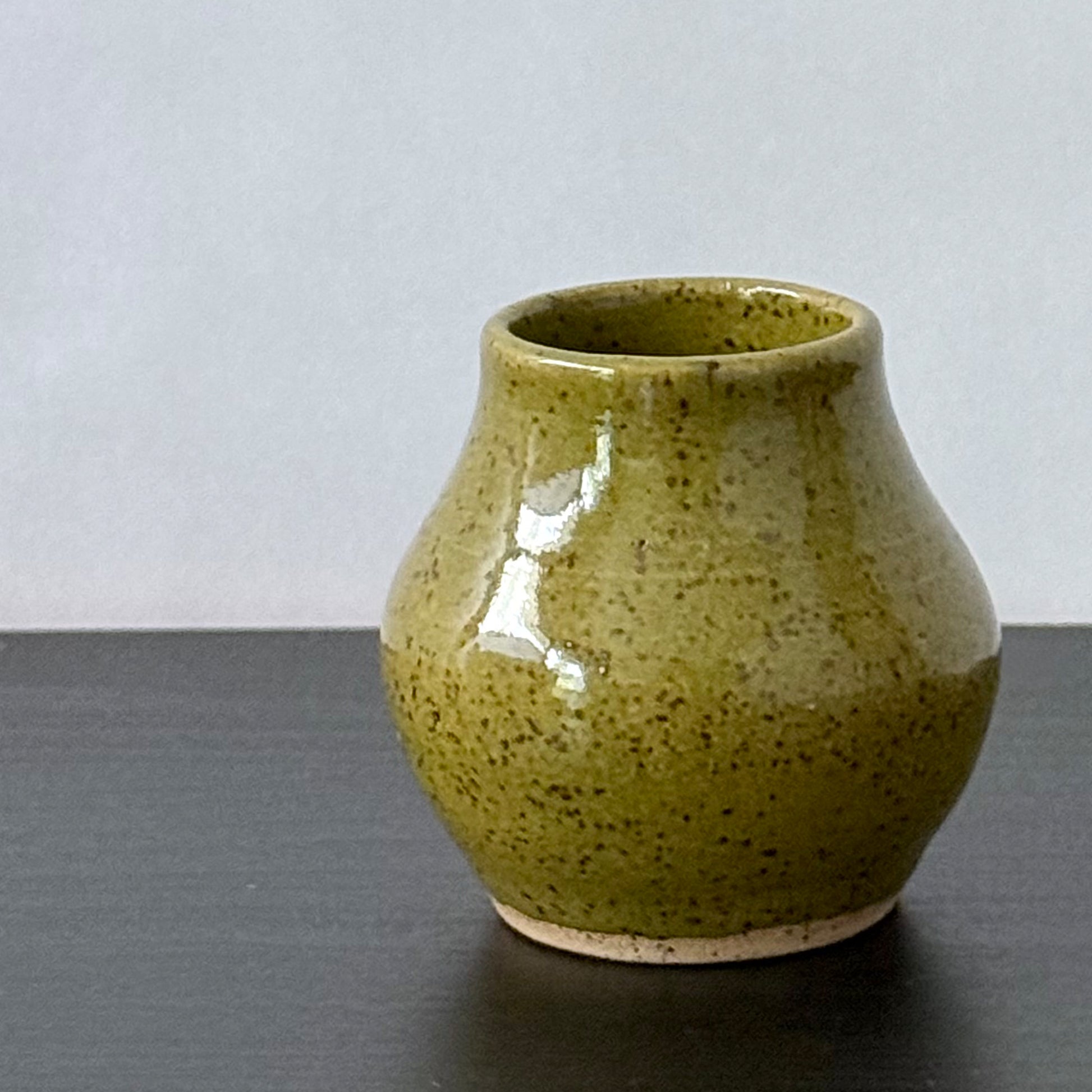 A small handmade ceramic vase with a rounded body and short flared rim, glazed in olive green with dark brown speckles, sitting on a dark surface against a light gray background.