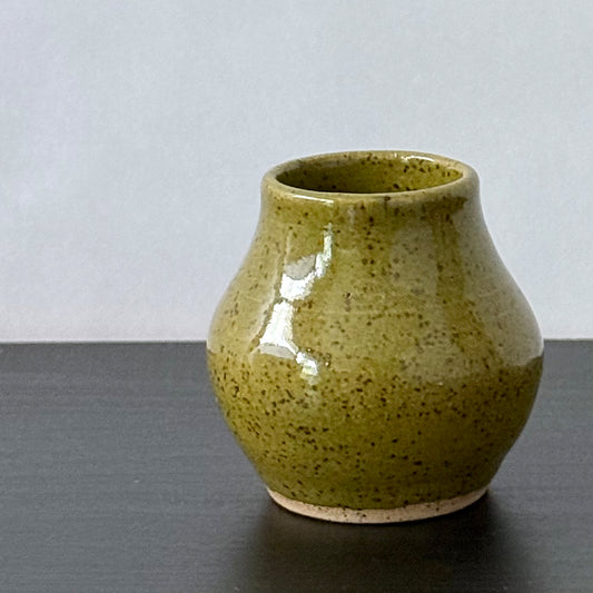 A small handmade ceramic vase with a rounded body and short flared rim, glazed in olive green with dark brown speckles, sitting on a dark surface against a light gray background.