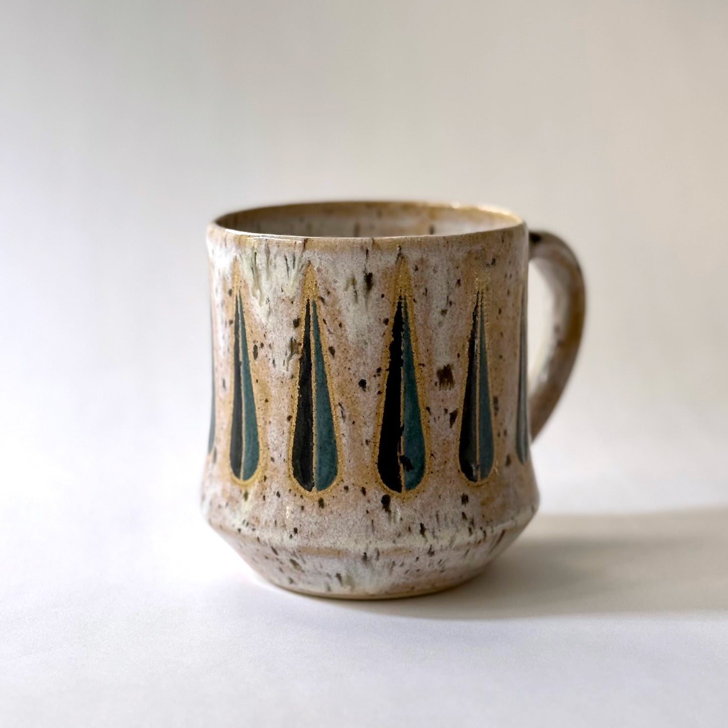 Handmade stoneware mug with a teal-and-black teardrop pattern on a speckled cream glaze, photographed against a white background.
