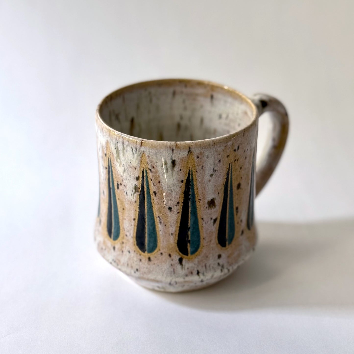 Handmade stoneware mug with a teal-and-black teardrop pattern on a speckled cream glaze, photographed against a white background.