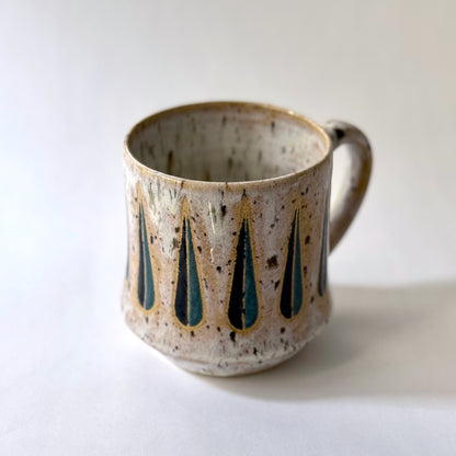Handmade stoneware mug with a teal-and-black teardrop pattern on a speckled cream glaze, photographed against a white background.