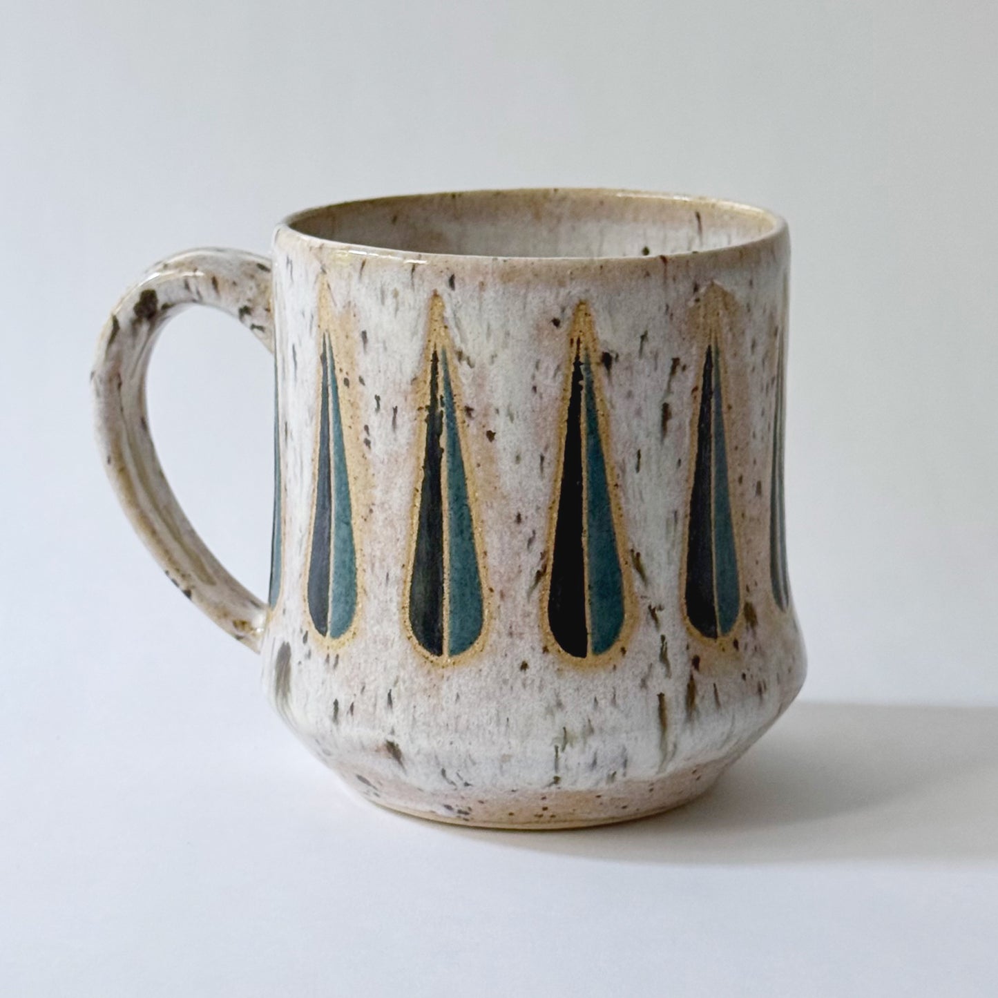 Handmade stoneware mug with a teal-and-black teardrop pattern on a speckled cream glaze, photographed against a white background.