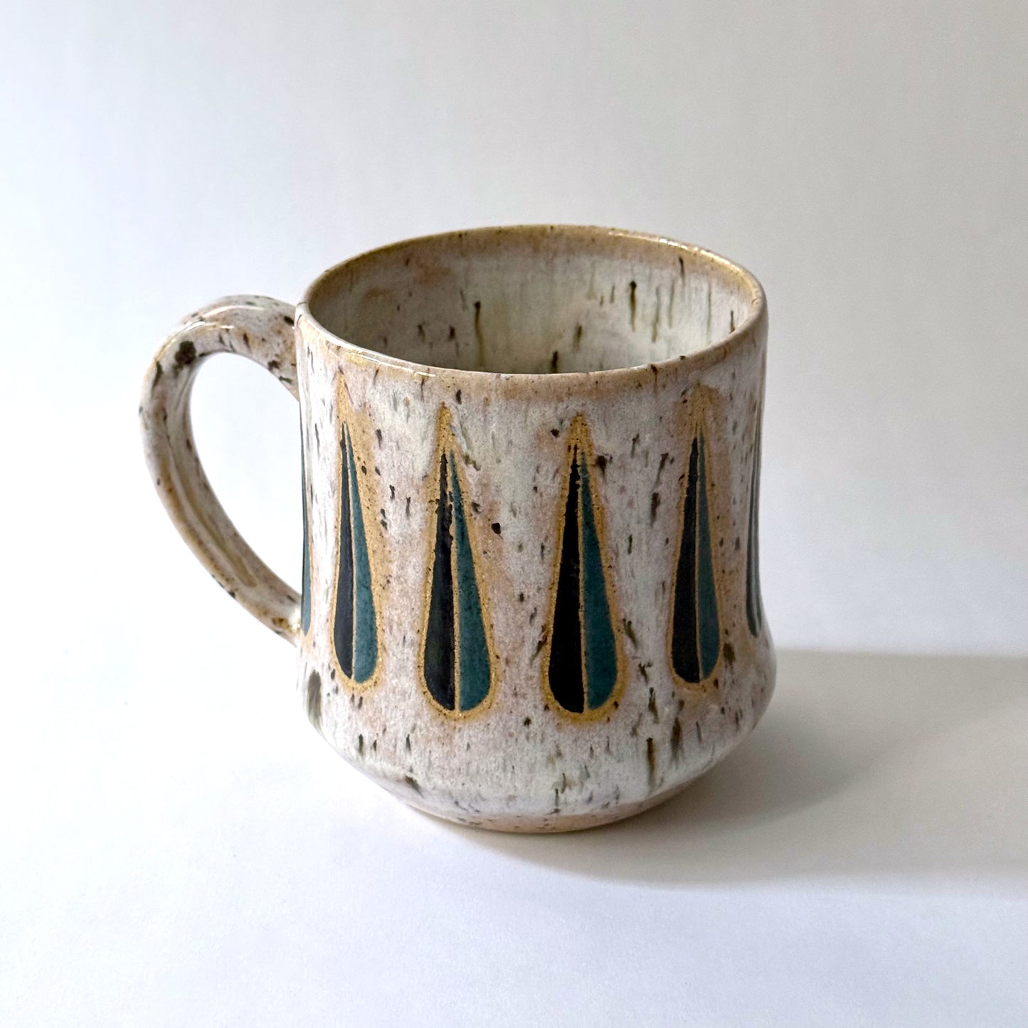 Handmade stoneware mug with a teal-and-black teardrop pattern on a speckled cream glaze, photographed against a white background.