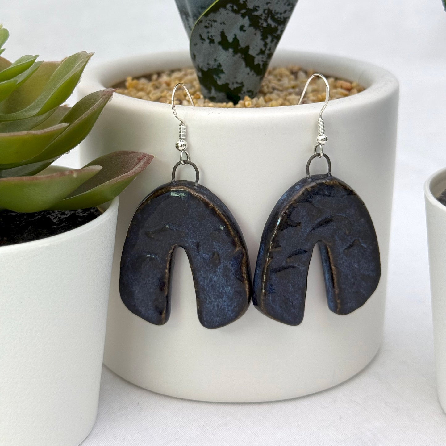 Pair of dark blue earrings hanging in front of a white pot with plants.