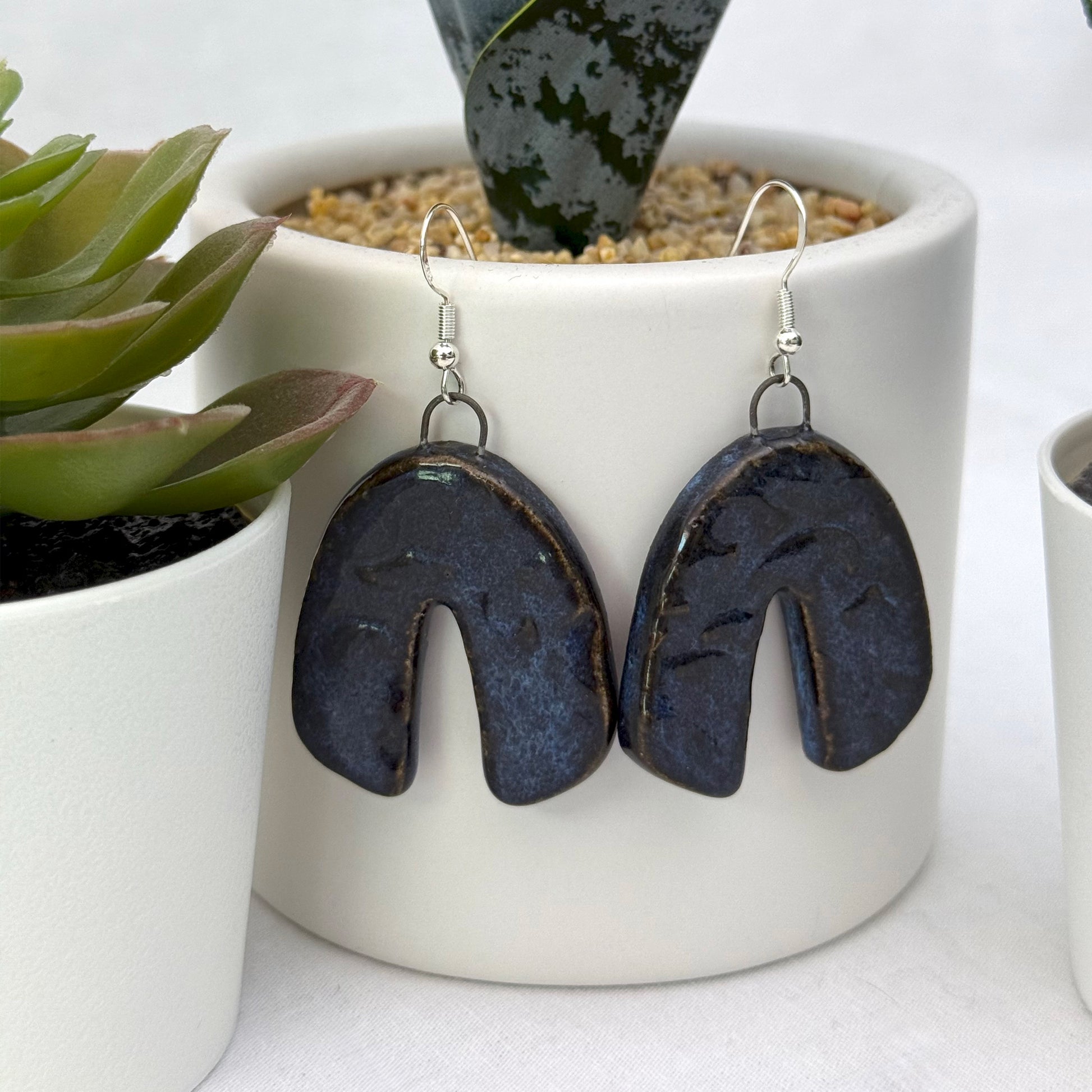 Pair of dark blue earrings hanging in front of a white pot with plants.
