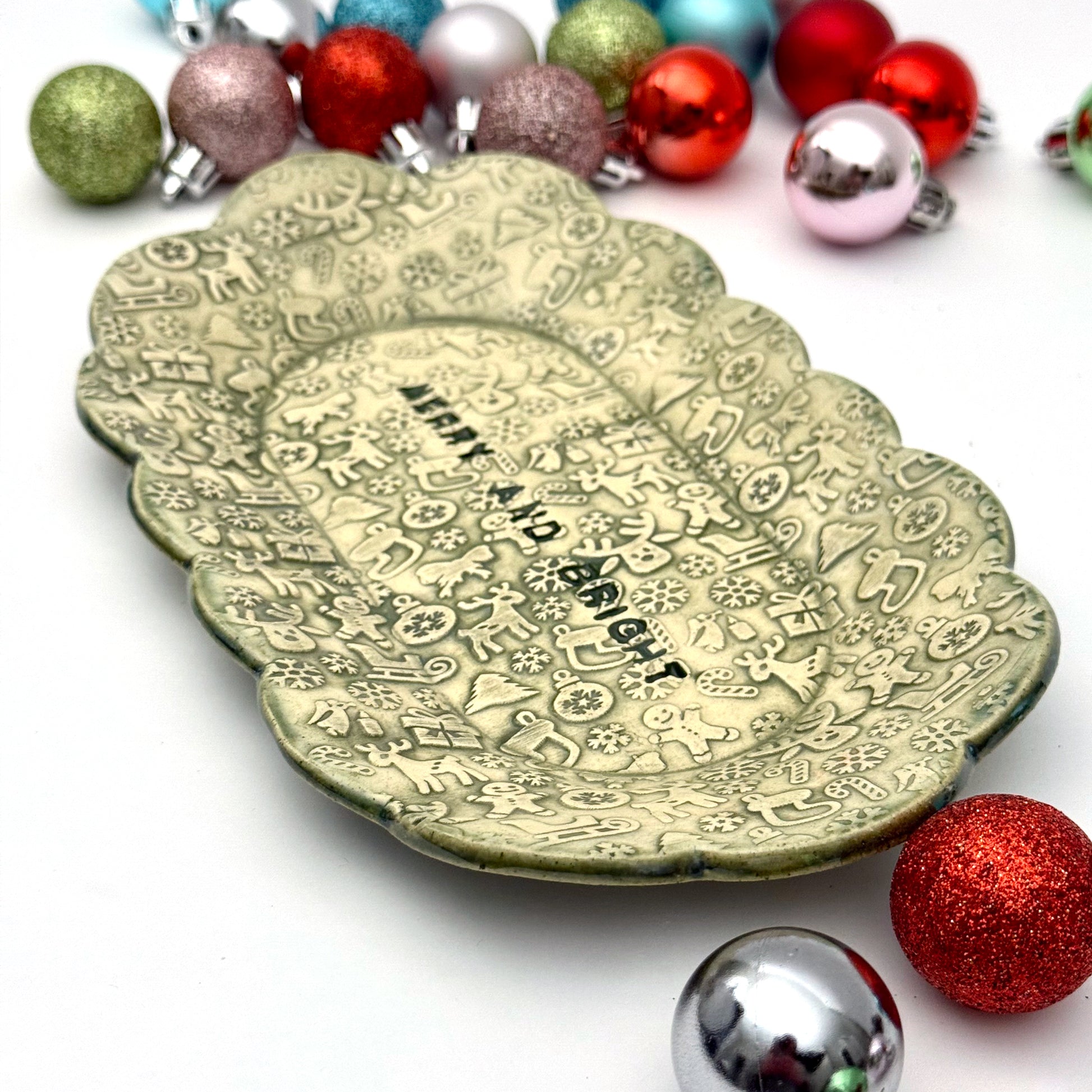 A green ceramic plate with scalloped edges and embossed holiday icons, featuring the phrase “MERRY AND BRIGHT” at the center, displayed among assorted ornaments.