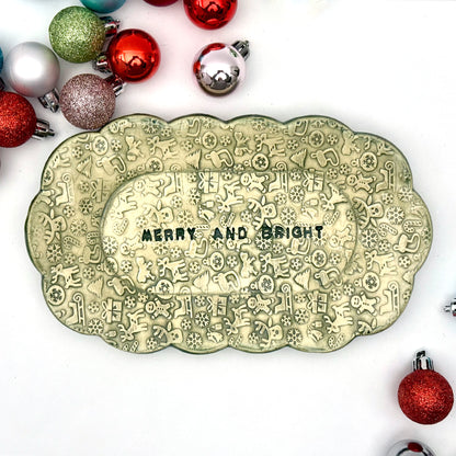 A green ceramic plate with scalloped edges and embossed holiday icons, featuring the phrase “MERRY AND BRIGHT” at the center, displayed among assorted ornaments.