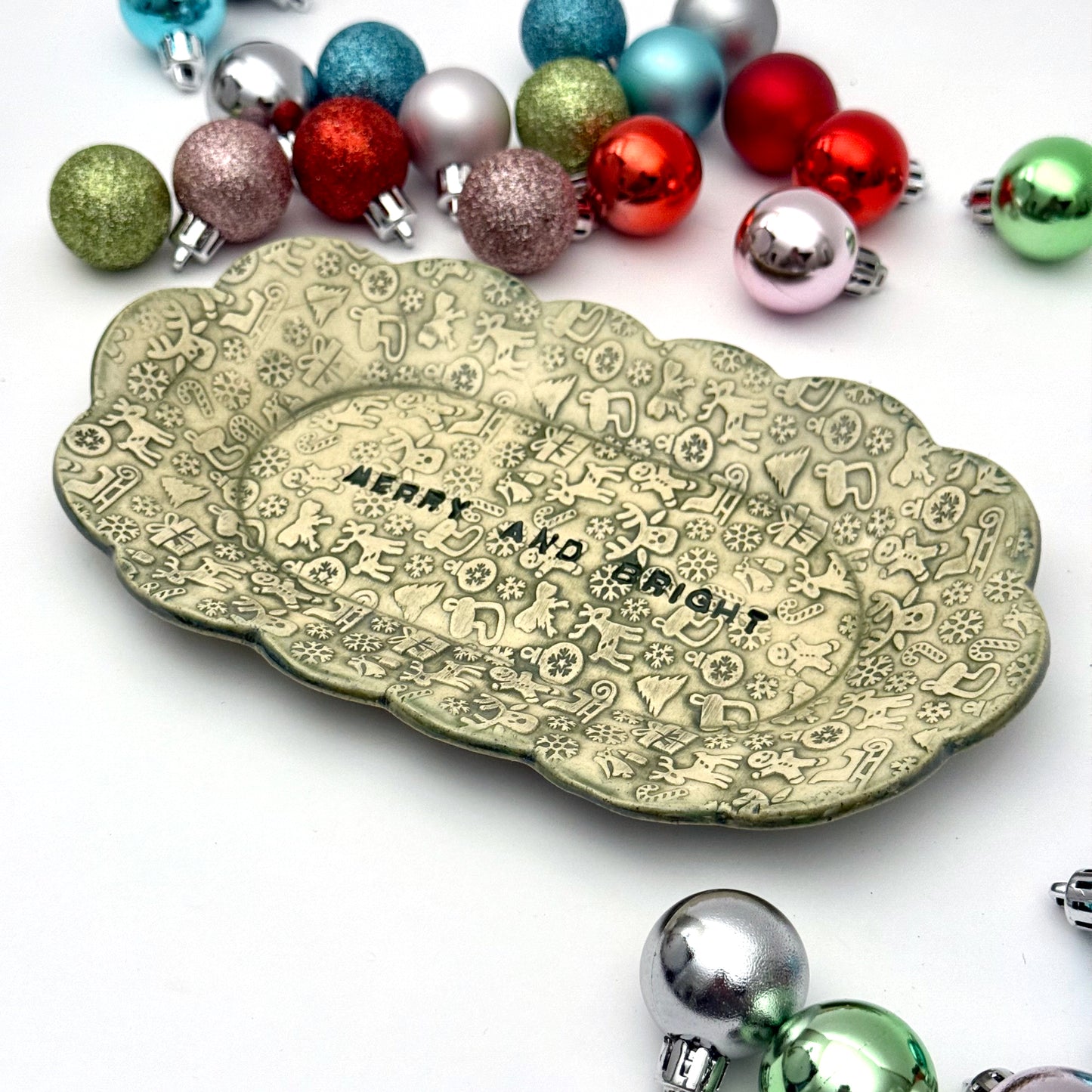 A green ceramic plate with scalloped edges and embossed holiday icons, featuring the phrase “MERRY AND BRIGHT” at the center, displayed among assorted ornaments.