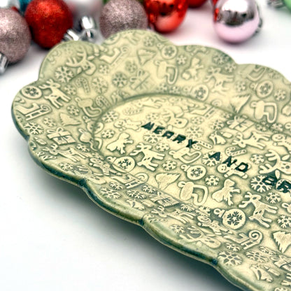 Close-up of a green ceramic plate with scalloped edges and embossed holiday icons, featuring the phrase “MERRY AND BRIGHT” at the center, displayed among assorted ornaments.