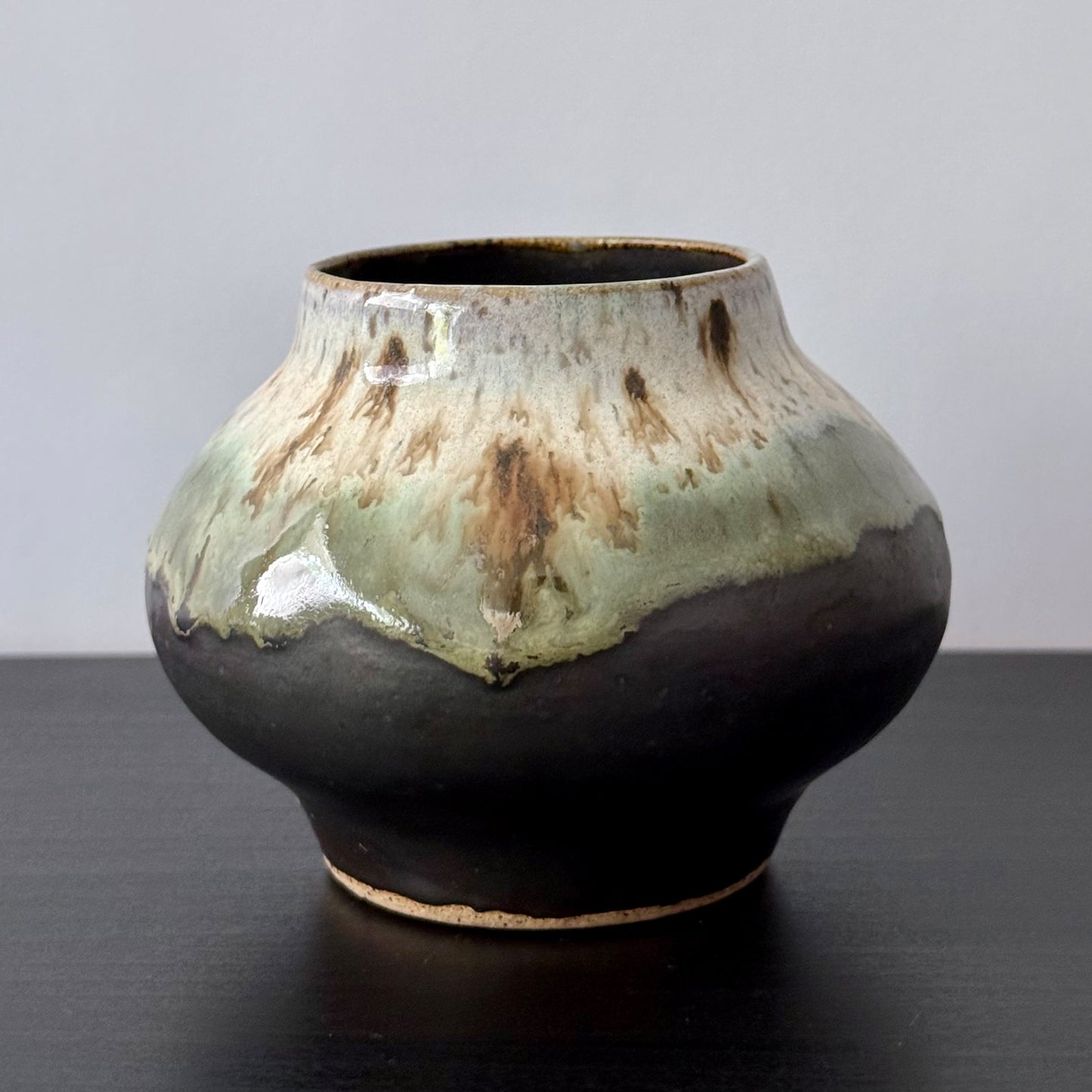 A handmade ceramic pot with a rounded bulbous body, a narrow flared neck, and a glossy glaze featuring a dark brown base with a light green and beige lava-like dripped pattern on the upper half, set against a neutral gray background and dark wooden surface.
