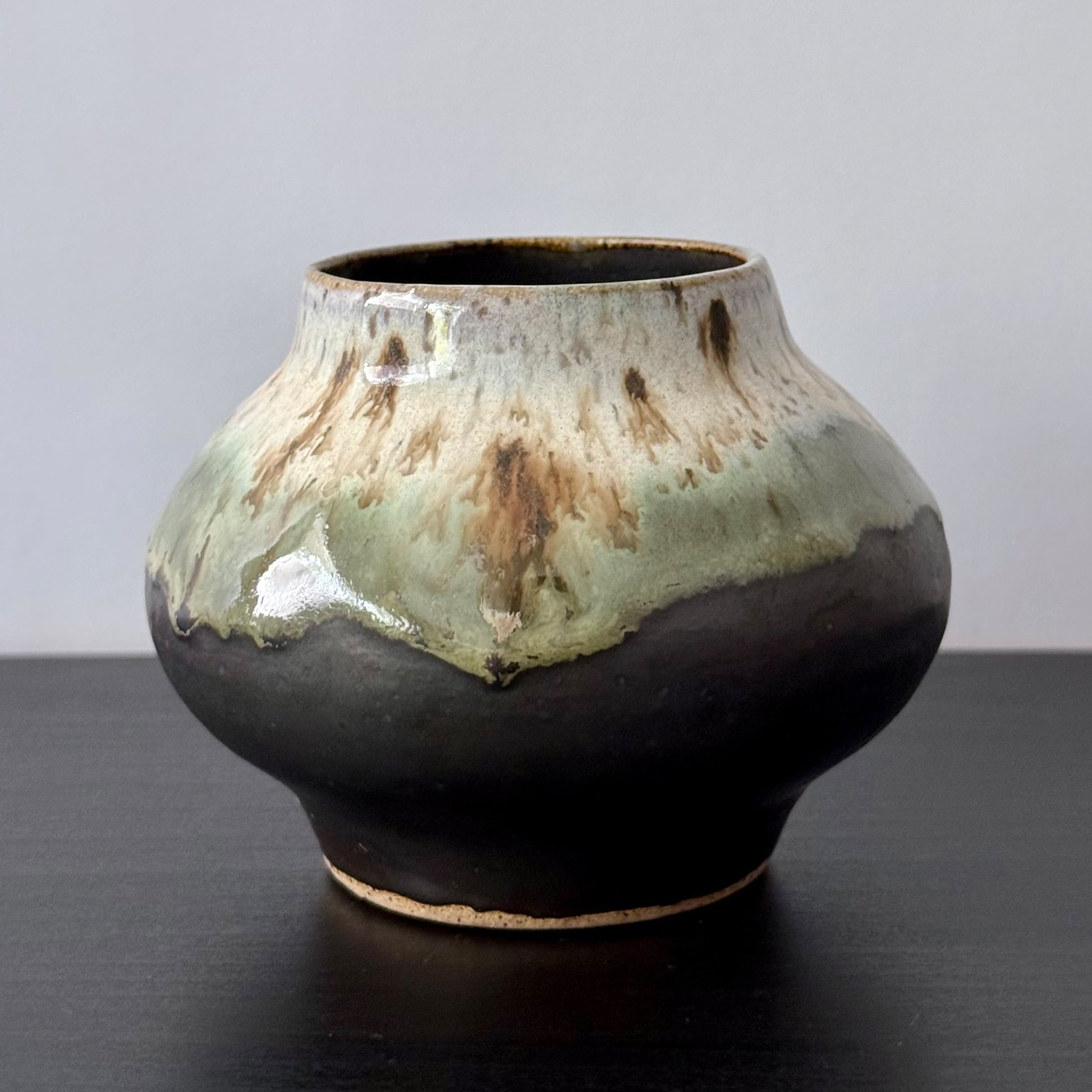 A handmade ceramic pot with a rounded bulbous body, a narrow flared neck, and a glossy glaze featuring a dark brown base with a light green and beige lava-like dripped pattern on the upper half, set against a neutral gray background and dark wooden surface.