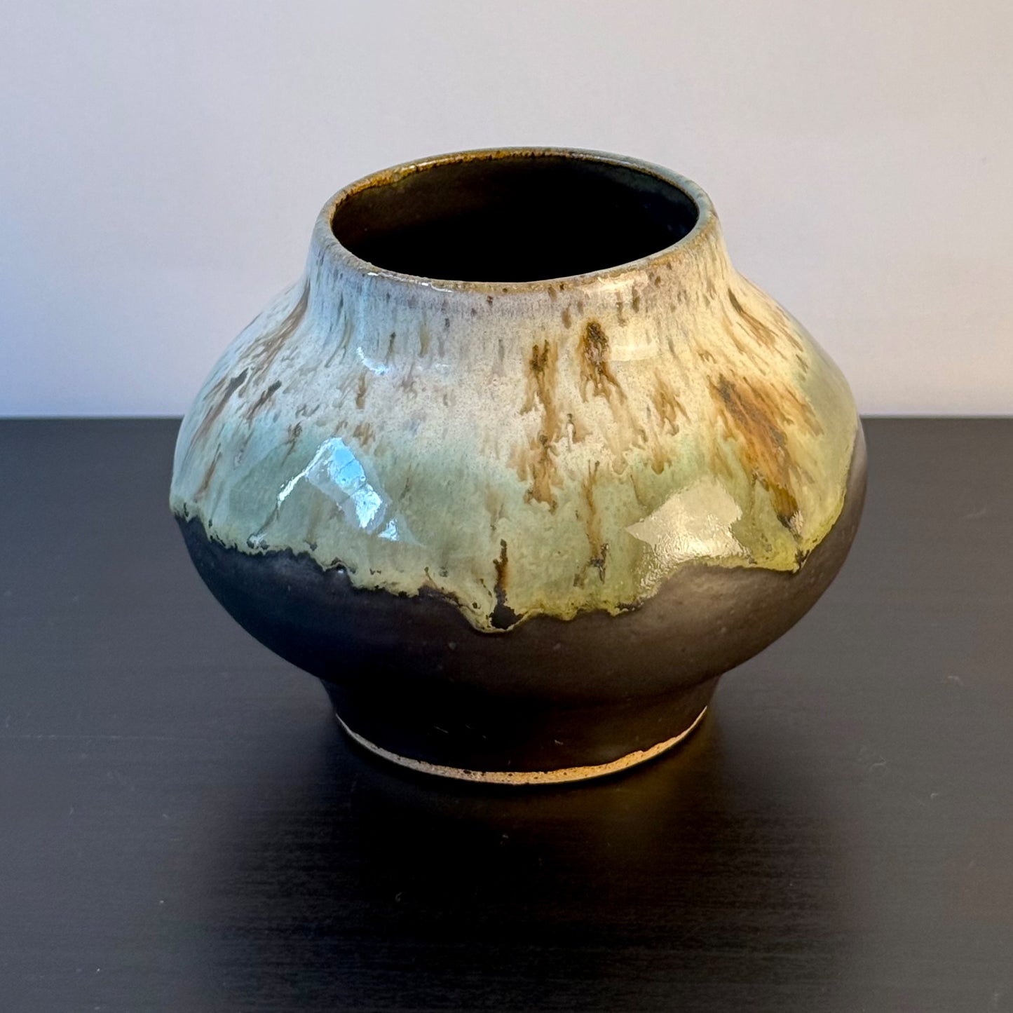 A handmade ceramic pot with a rounded body, a flared rim, and a glossy glaze featuring a dark brown base with a dripping light green and beige glaze on top, set on a dark table against a light gray background.