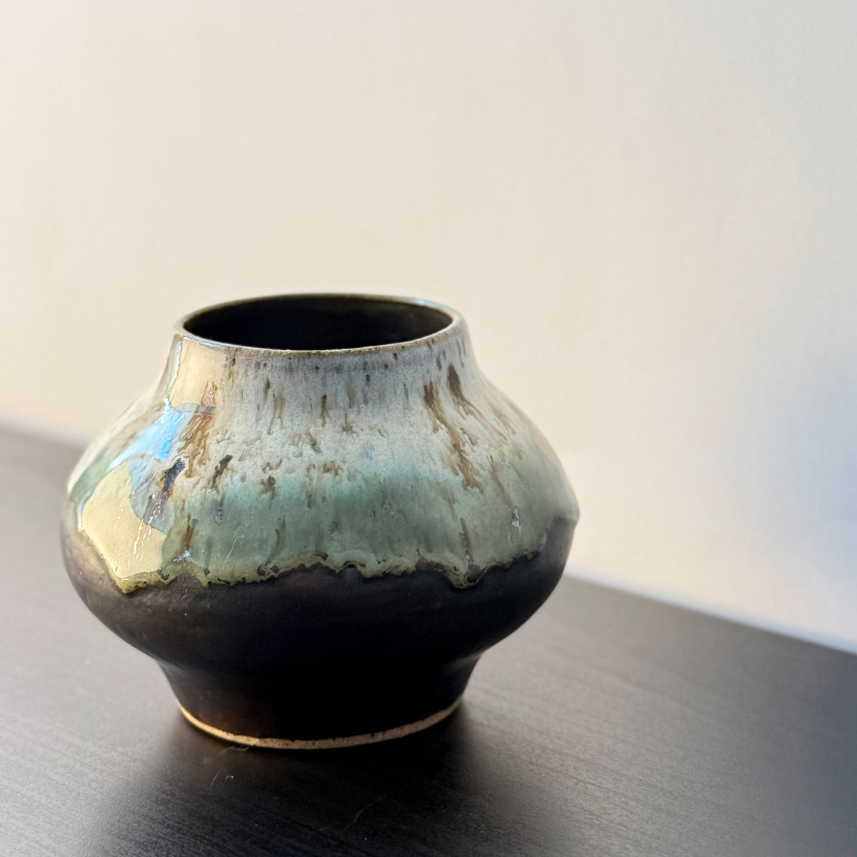 A small handmade ceramic pot with a rounded bulbous body, featuring a glossy dark brown base and a light blue-green glaze dripping over the top, set on a dark wooden surface against a plain light background.