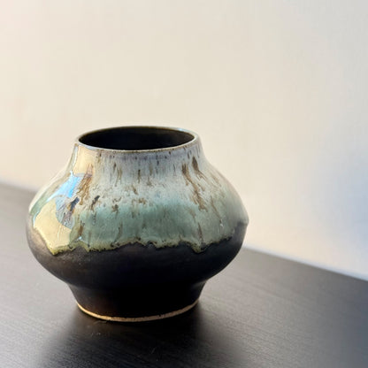 A small handmade ceramic pot with a rounded bulbous body, featuring a glossy dark brown base and a light blue-green glaze dripping over the top, set on a dark wooden surface against a plain light background.