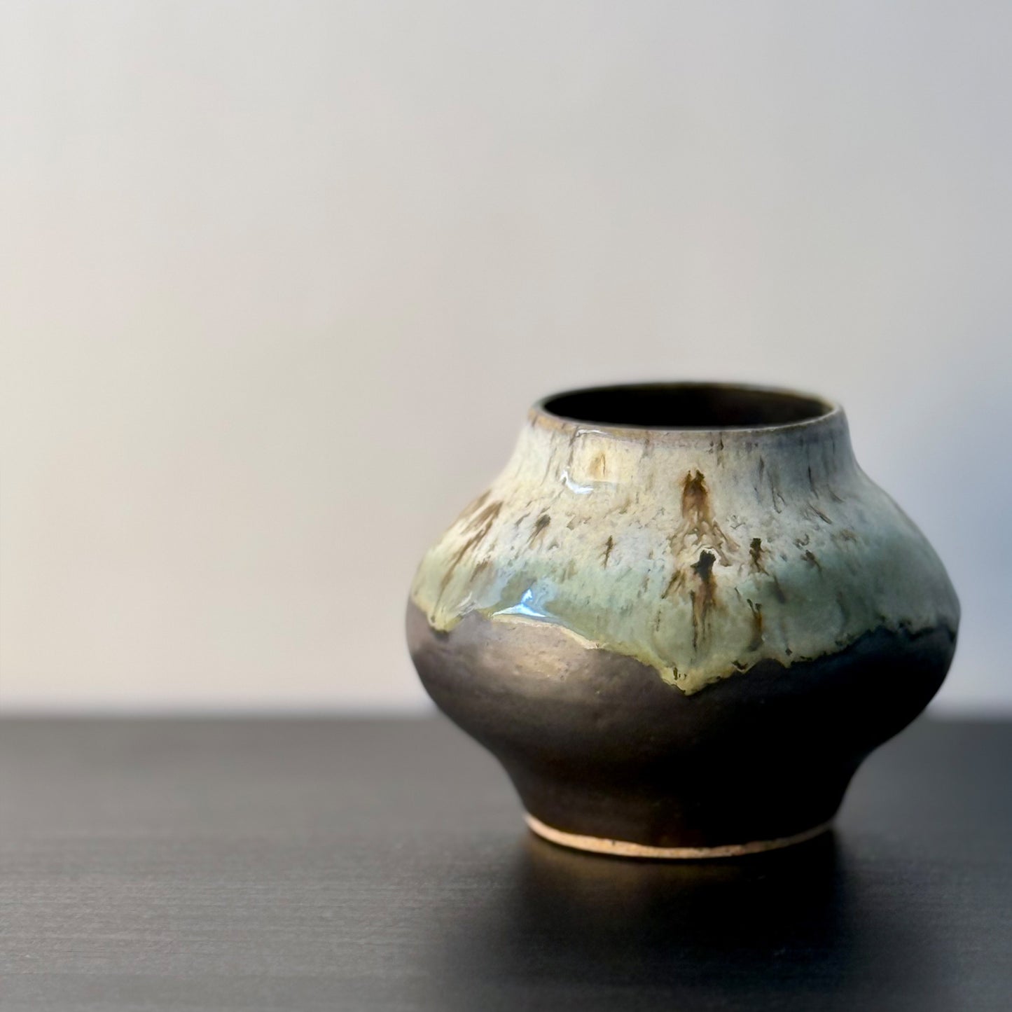 A small handmade ceramic pot with a rounded bulbous body, featuring a glossy glaze that transitions from pale sage green and cream at the top to a dark charcoal-brown at the base, sitting on a dark surface against a neutral light gray background.