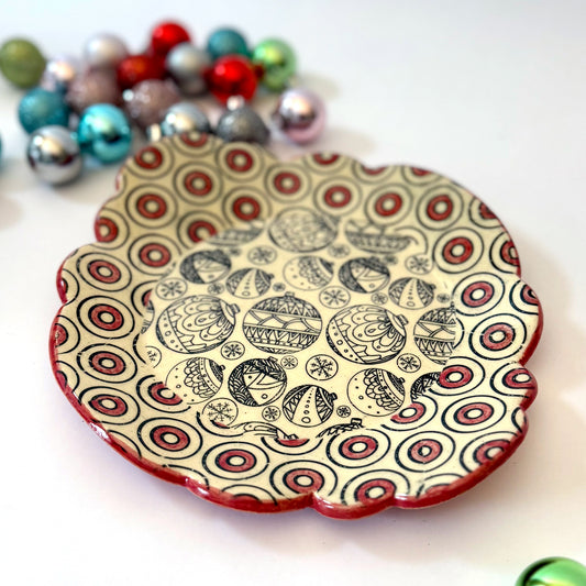 Scalloped plate with black holiday ornament designs and a red-and-black circle border, shown with colorful ornaments in the background.