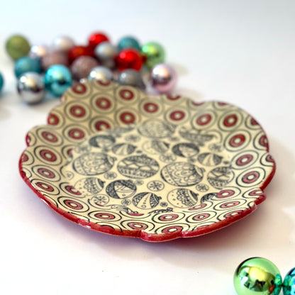 Scalloped plate with black holiday ornament designs and a red-and-black circle border, shown with colorful ornaments in the background.