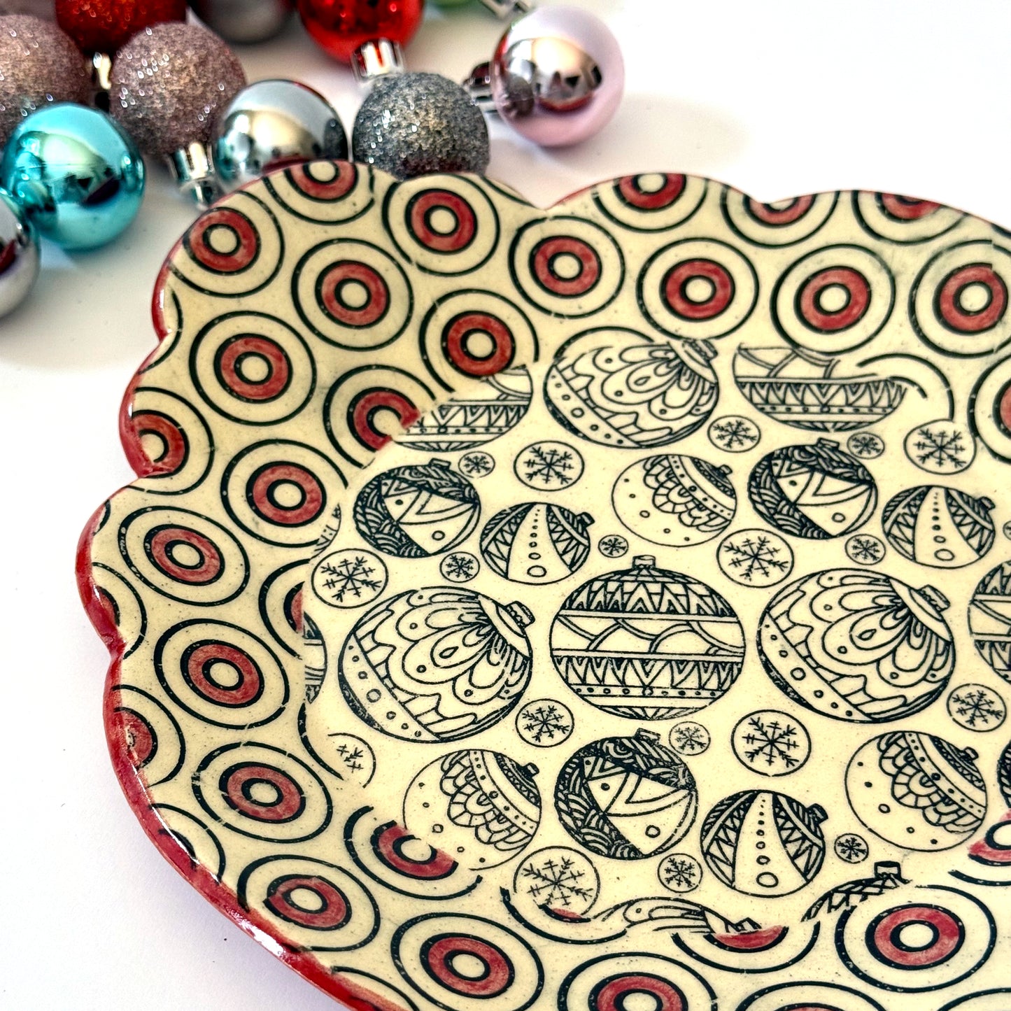 Scalloped plate with black holiday ornament designs and a red-and-black circle border, shown with colorful ornaments in the background.
