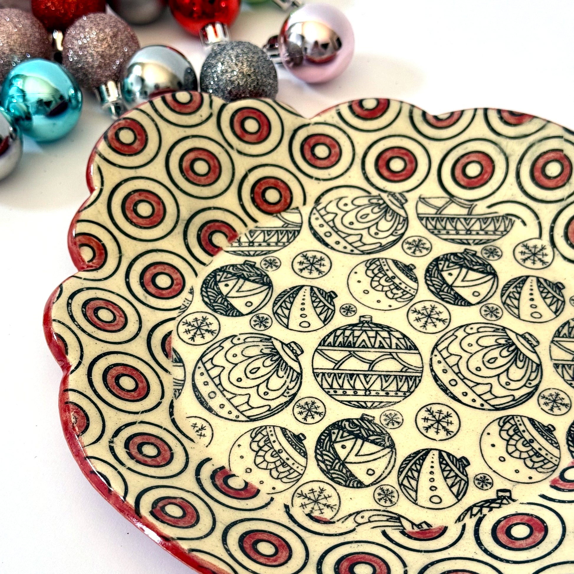Scalloped plate with black holiday ornament designs and a red-and-black circle border, shown with colorful ornaments in the background.