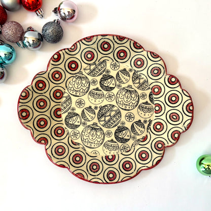 Scalloped plate with black holiday ornament designs and a red-and-black circle border, shown with colorful ornaments in the background.