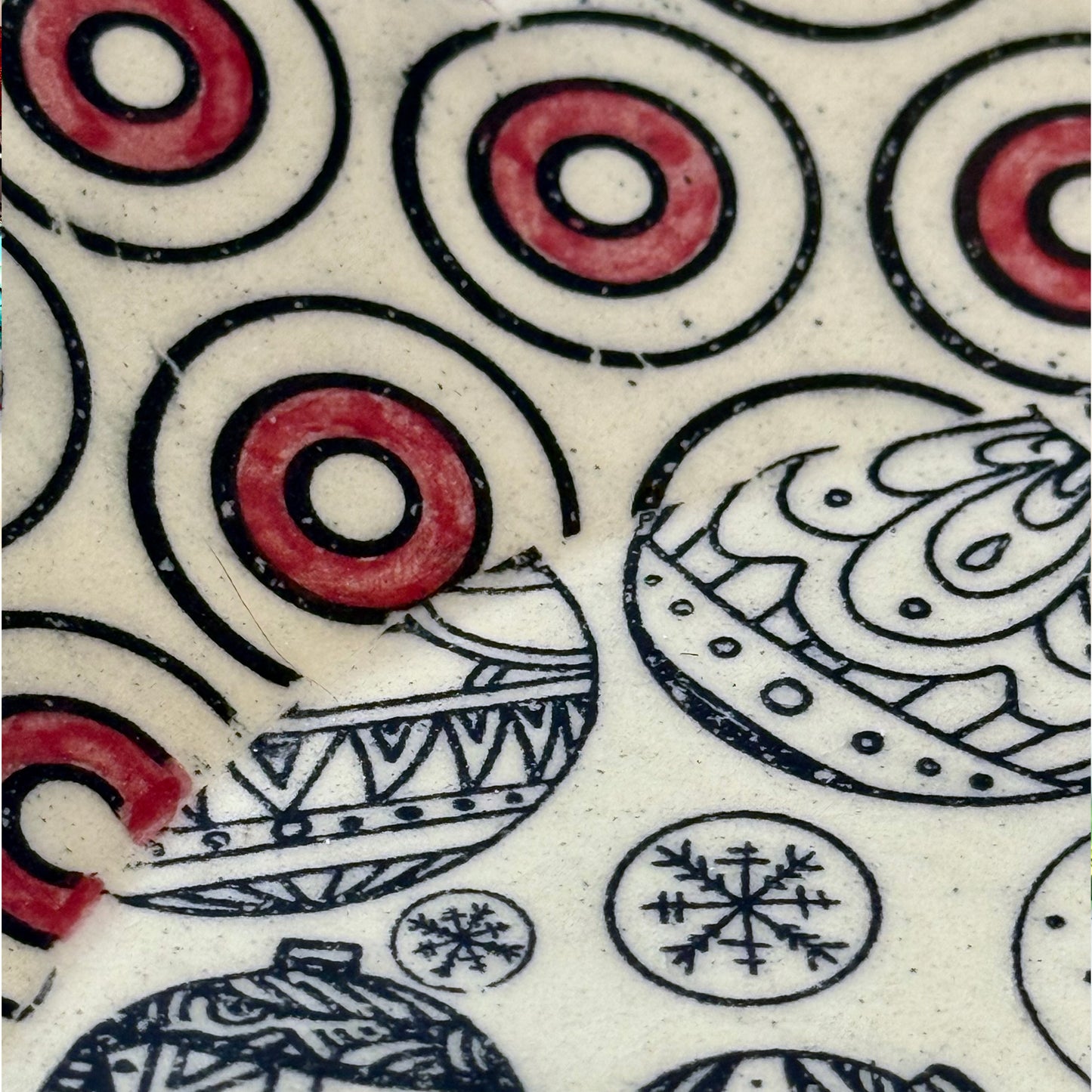 Close-up of scalloped plate with black holiday ornament designs and a red-and-black circle border, shown with colorful ornaments in the background.