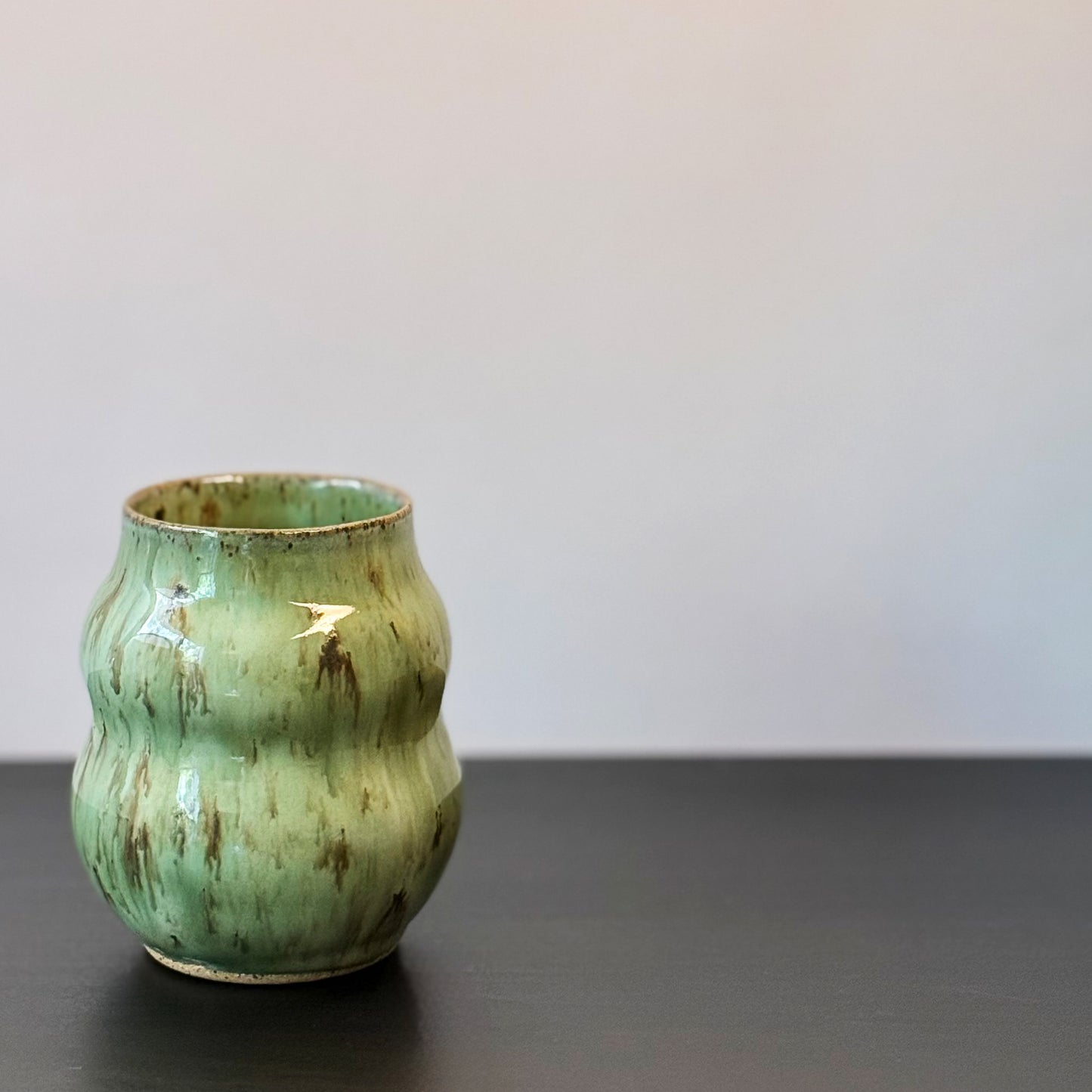 Handmade ceramic vase with a textured, ribbed body and a glossy light green glaze with brown distressed accents, sitting on a dark surface against a plain light gray wall.