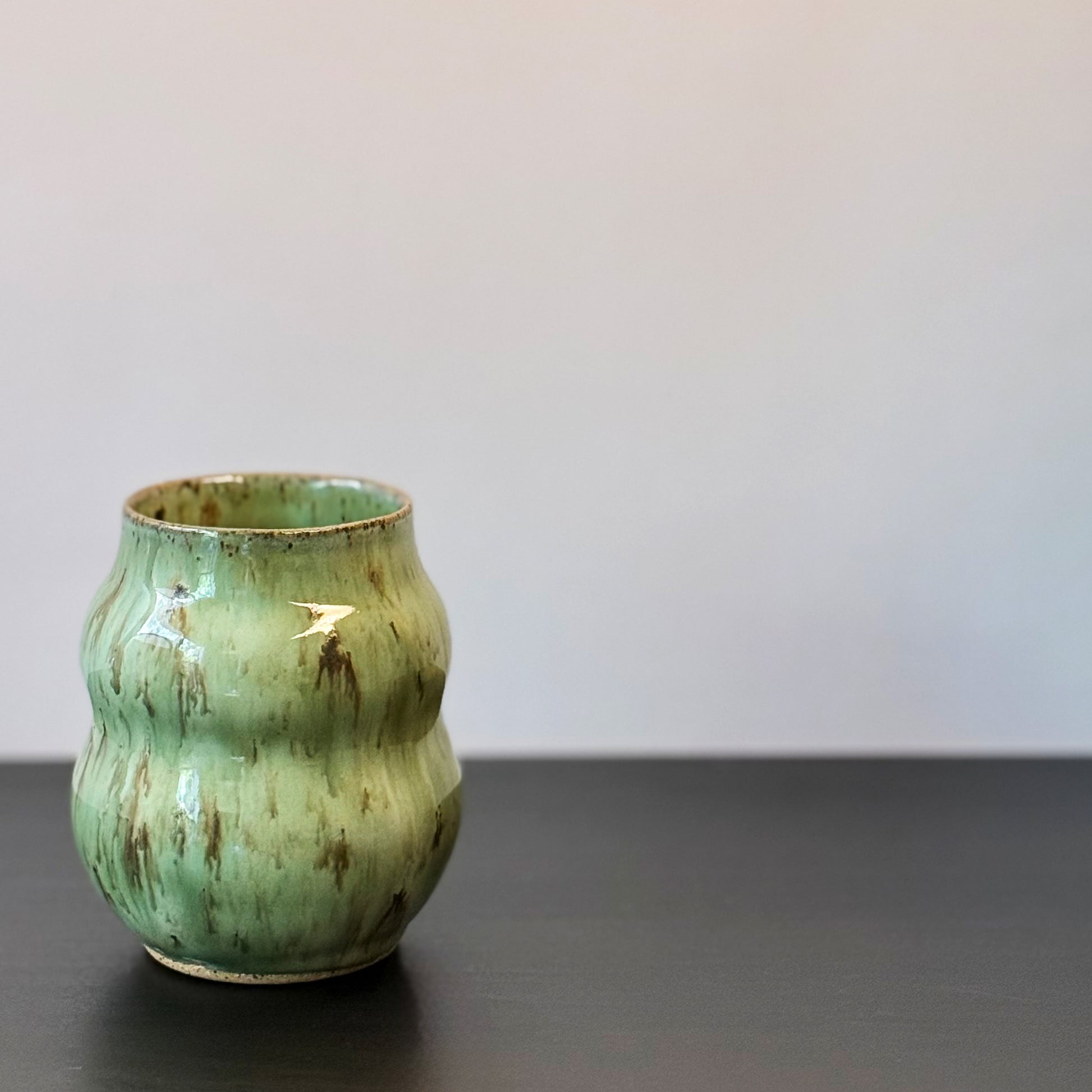 Handmade ceramic vase with a textured, ribbed body and a glossy light green glaze with brown distressed accents, sitting on a dark surface against a plain light gray wall.