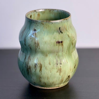 Handmade ceramic vase with a textured, ribbed body and a glossy light green glaze with brown distressed accents, sitting on a dark surface against a plain light gray wall.